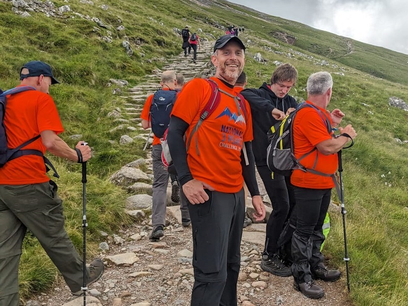 man-turns-to-smile-at-camera-as-team-walk-up-mountain-path-behind-him